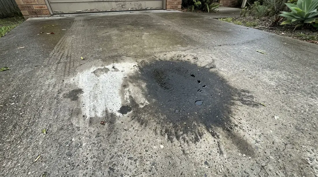 A dark engine oil stain on a broom-finished concrete driveway with degreaser being brushed into the surface